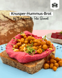 Eine Scheibe Brot auf einem Teller mit pinkfarbenem Rote-Beete-Hummus und Kichererbsen. Im Hintergrund ein Laib Brot.